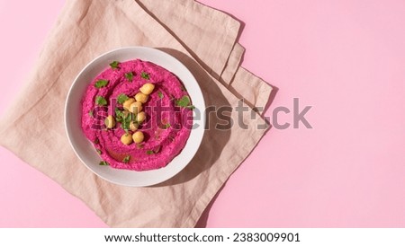 White plate of beetroot hummus decorated with chickpeas on pink towel and background with copy space, top view, hard shadow
