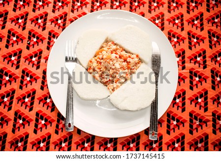 A white plate of boiled yam and fried scrambled eggs prepared with sliced onions, tomatoes and red pepper. Served on a colorful red pattern tablecloth with a knife and fork  placed at the side