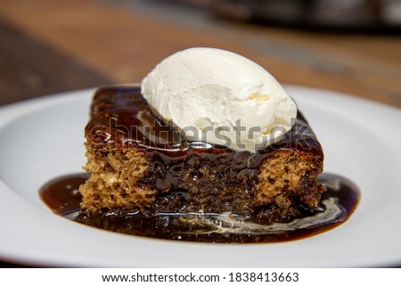A white plate of delicious sticky toffee pudding chocolate cake with cream and chocolate sauce on a wooden kitchen work top
