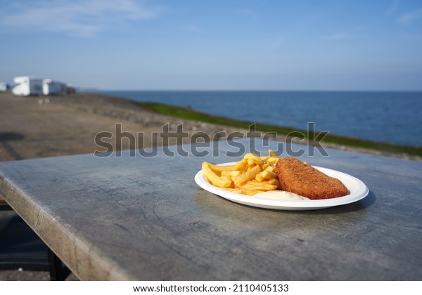 White plate with fast food on the\
beach. Schnitzel and fries with sauce on the table. Sea water and\
mobile homes in the deep blur. South Holland,\
Brouwersdam.