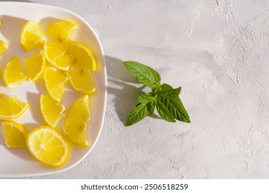 A white plate filled with lemon wedges sits next to a sprig of fresh mint on a white surface - Powered by Shutterstock