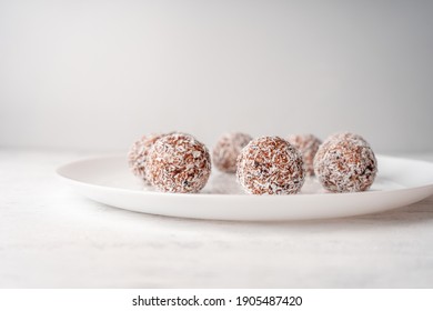 A white plate with round sweets made of dates and coconut shavings on a light wooden background.