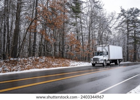 White popular middle duty white day cab rig semi truck with box trailer for local freights and deliveries driving on the winter slippery road with snow and ice and snowy forest on the side