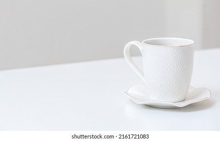 White Porcelain Cup On A Table Indoors. White Saucer And Cup Of Coffee On White Table. Ceramic Tea Cup On White Kitchen Table. Side View