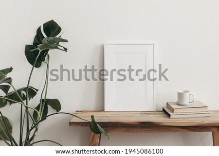 White portrait frame mockups on vintage bench, table. Cup of coffee on pile of books and monstera potted plant. White wall background. Scandinavian interior, neutral color palette. Artistic display.