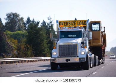 White Powerful Big Rig Semi Truck With Oversize Load Sign And Step Down Trailer Carry And Transporting Large Oversize Digger On Multiline Wide Highway