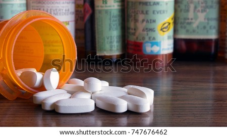 White prescription pills spilled onto a table with many prescription bottles in the background. Concepts of opioid addiction and doctor shopping for prescription pain killers