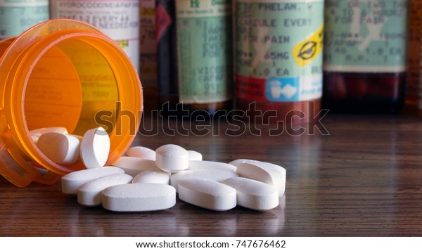 White prescription\
pills spilled onto a table with many prescription bottles in the\
background. Concepts of opioid addiction and doctor shopping for\
prescription pain killers