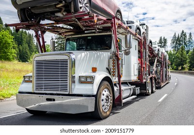 White professional industrial big rig car hauler semi truck transporting cars on the two level modular semi trailer running on the road with green mountains of summer Oregon on the background