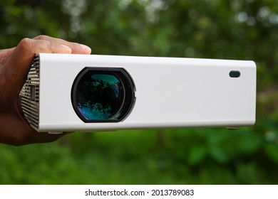 A White Projector With Green Shiny Lens In Hands At Outdoor, It's A Chinese LED Projector Front Face.