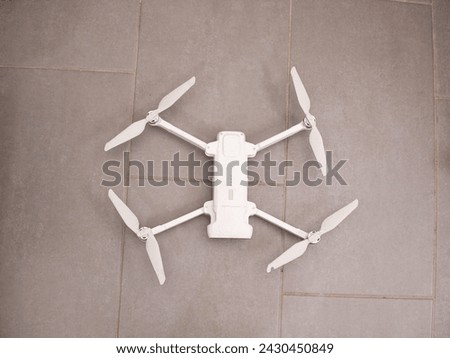 A white quadcopter is parked on top of a tile floor. The drone appears stationary, resting on the flat surface of the floor tiles.