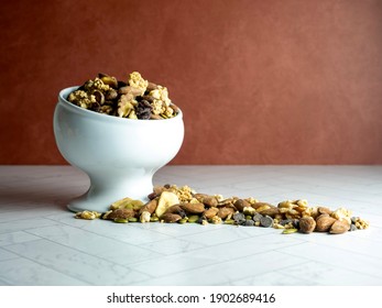 White raised bowl fill of trail mix, nuts and dried fruit spilling out onto the herringbone counter with a terra-cotta wall behind.  Peanuts, granola, pecans, walnuts, banana slices, cranberry. - Powered by Shutterstock