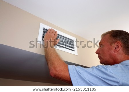 White rectangle air vent on a beige wall with a males hand on left side of the outflow furnace register. Mature male with left hand on and inspecting white home furnace air duct vent in a modern home