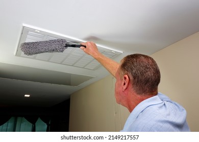 White rectangle HVAC air intake vent being dusted with a gray microfiber cloth dusting wand by an adult male. Caucasian male in his 50s dusting a ceiling intake HVAC furnace vent with a grey duster