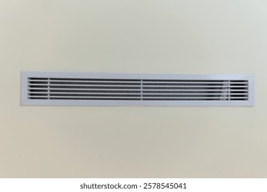 a white rectangular air vent set against a plain, light beige wall. The vent is characterized by its horizontal slats. It's a simple, minimalist image of a common architectural feature.