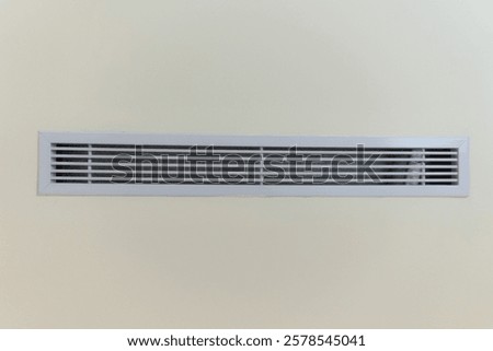 a white rectangular air vent set against a plain, light beige wall. The vent is characterized by its horizontal slats. It's a simple, minimalist image of a common architectural feature.