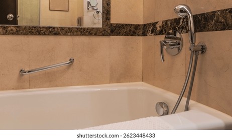 A white rectangular bathtub in the corner of the bathroom. A towel on the edge. Taps, a shower head, and a metal handrail are mounted on the beige tiled wall. Reflection in the wall mirror.
