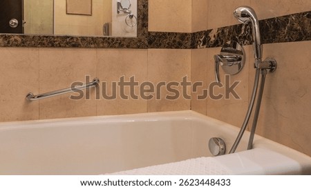 A white rectangular bathtub in the corner of the bathroom. A towel on the edge. Taps, a shower head, and a metal handrail are mounted on the beige tiled wall. Reflection in the wall mirror.
