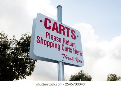White rectangular Please Return Shopping Carts Here sign at a parking lot of a store or supermarket marking a place where shopping carts should be returned