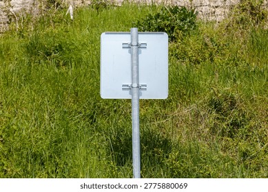 White rectangular sign stands in tall green grass. Metal pole supports blank sign. Sign shows no text or symbols. Background includes stone wall and weeds. Grass grows around sign