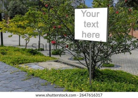 A white rectangular signboard mounted on a metal post, placed in a lush green park setting with leafy trees in the background.