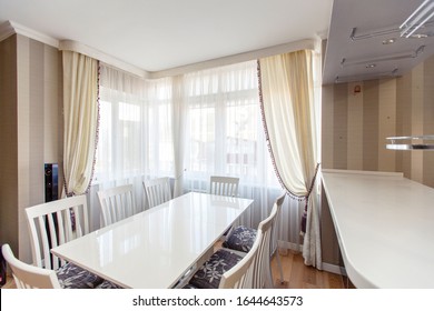 White rectangular table with white high-backed chairs in the kitchen-living room of the laksheri cottage in classic style. Eight chairs, large Windows with white tulle curtains