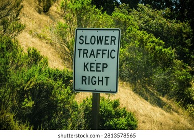 A white rectangular traffic sign with black lettering instructing drivers to keep right for slower vehicles is mounted on a wooden post amidst lush green foliage and dry grass on a hillside