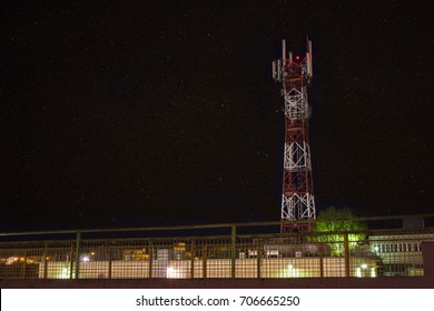 White And Red Cell Tower With Attached Antennas. In The Background A Night With Starry Sky.