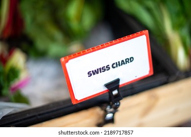 A White And Red Colored Swiss Chard Sign With Black Text Attached To A Small Wooden Crate Containing Green Leafy Swiss Chard. The Produce Card Is Attached With A Small Black Plastic Clamp. 