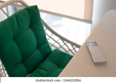 White Remote Control Panel For Automatic Roller Shades Lies On A Beige Table In The Living Room. Nearby Is A Chair With A Green Pillow. On The Window, A Roller Blind With Automatic Control. 