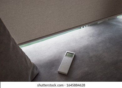 White Remote Control Panel For Motorized Roller Shades. Automatic Roller Blinds Beige Color On The Window. Remote Control Shades Are Near The Sofa With The Pillow. Sofa Covered With Pastel Velours.