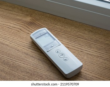 White Remote Control Panel For Motorized Roller Shades Or Automatic Blinds Lies On A Wooden Windowsill Near The Window.