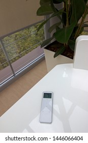 White Remote Control Panel For Roller Blinds, Which Lies On A White Table Near Window With Beige Automatic Roller Shades.