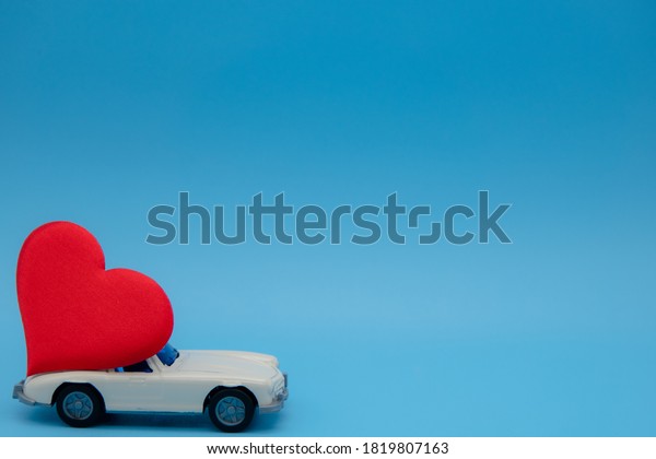 White retro toy car\
delivering a heart on a blue background. Postcard February 14,\
Valentine\'s Day. Flower delivery. March 8, International Happy\
Women\'s Day.