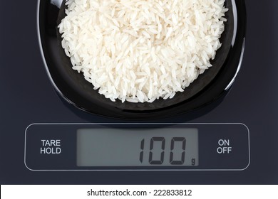 White Rice In A Black Plate On Digital Scale Displaying 100 Gram.