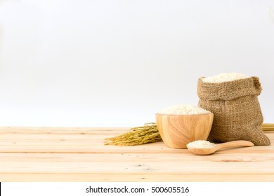 White Rice In Bowl And A Bag, A Wooden Spoon And Rice Plant On A Wooden Table, Isolated In A White Background, Side View With Copy Space