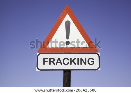 White road warning triangle with black  exclamation point and red frame on  a wooden mast in front of a blue sky. A second rectangular sign warns in english about fracking