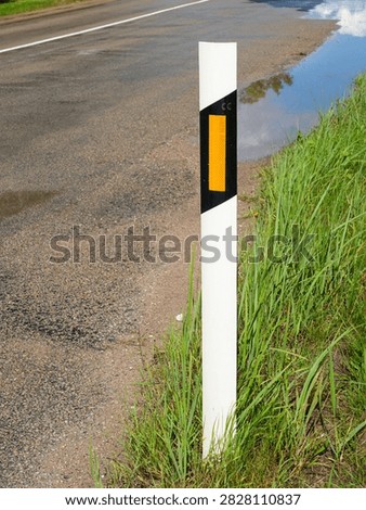 White roadside delineator post with orange reflector stands beside wet asphalt road, marking the road edge for improved driver visibility and safety