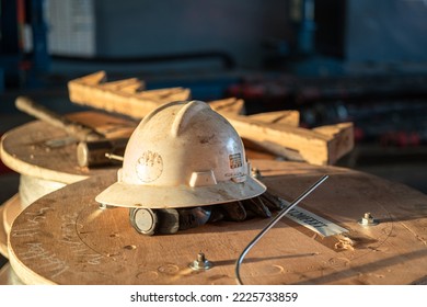 A White Safety Working Hardhat Or Helmet In Orange Sun Lighting Which Is Placed On The Material Wooden Reel Ac The Factory. Industrial Safety Equipment Object. Close-up And Selective Focus.