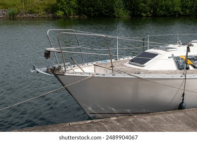 A white sailboat is docked at a marina, with a wooden dock and calm water in the background. The boat is tied to the dock with ropes, and the bow is facing towards the camera. - Powered by Shutterstock