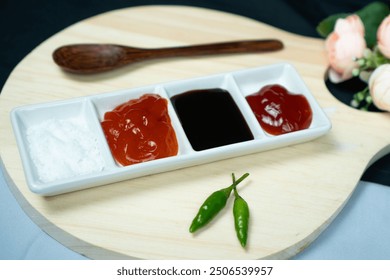 White sauce plate containing ketchup, chili sauce, sweet soy sauce and salt, with green chili and wooden spoon on a wooden tray with flower - Powered by Shutterstock