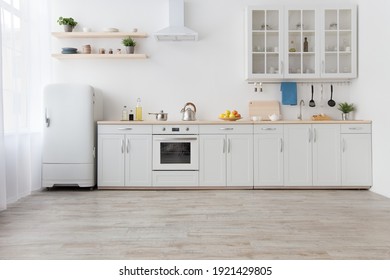White scandinavian kitchen interior with dining room and wooden floor. Simply furniture with utensils, shelves with crockery and plants in pot, refrigerator in modern minimal design, empty space - Shutterstock ID 1921429805