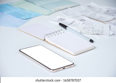 White Screen Of Mobile Phone Or Smartphone With Mini Note Book, A Black Pen, Utility Bills Or Pay Slips And Bank Deposit Account Booklets On White Table Blurred Background