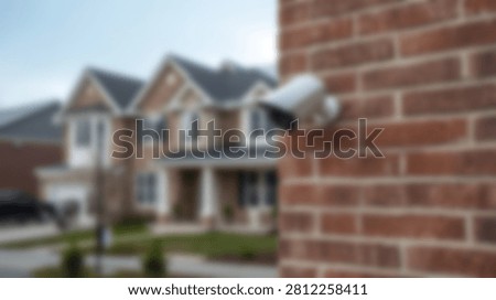 A white security camera is attached to a red brick wall, with a blurred residential house visible in the background.