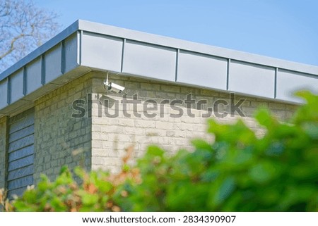 A white security camera is installed on the corner of a beige brick building with a metal roof edge, surrounded by greenery and set against a clear blue sky.