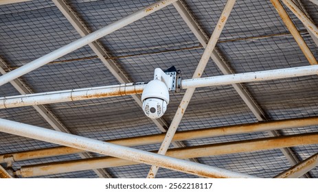 white security camera, mounted on a rusty metal pole, hangs from a ceiling grid.