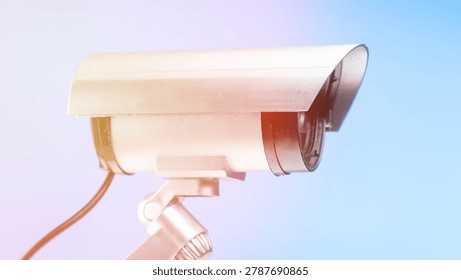 White security camera mounted on a stand. Close-up view shows metal casing and lens. Background features soft blue to purple gradient. Black wire connects to the camera base