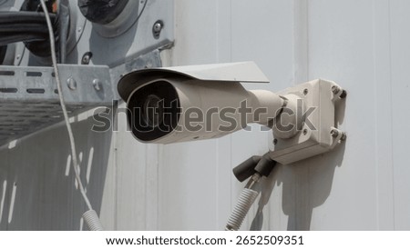 A white security surveillance camera mounted on a light-colored corrugated metal wall.