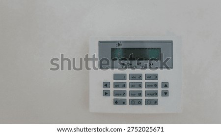 A white security system keypad mounted on a wall, featuring a small LCD screen and tactile buttons for arming and disarming
