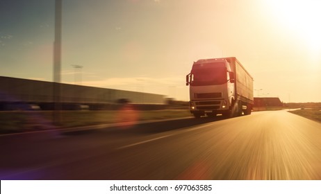 White Semi Truck With Cargo Trailer Attached Drives On The Empty Road. Industrial Warehouses By The Sides Of The Highway. Sunset. Blur Motion.